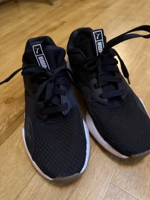 Svarta sneakers från Puma - Snygga svarta sneakers från Puma med vit sula och klassisk logga på plösen. Ovandelen är i mesh och syntetmaterial för bra andningsförmåga och komfort. Skorna har snörning och en sportig, modern look som passar till det mesta.