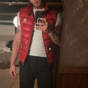 Röd dunväst från Moncler - Säljer en snygg röd dunväst från Moncler med quiltad design och hög krage. Västen har silverfärgade dragkedjor och Moncler-logga på bröstet. Perfekt att slänga över en t-shirt för en cool look. Kvitto finns från secondplace i Malmö 