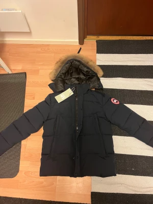 Mörkblå dunjacka Canada Goose M - Säljer en mörkblå dunjacka från Canada Goose med pälskantad huva och klassisk logga på ärmen. Hör av er vid minsta lilla fundering! Pris ej huggit i sten!