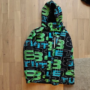 Färgglad vinterjacka från 8848 Altitude - Säljer en snygg vinterjacka från 8848 Altitude med coolt grafiskt mönster i grönt, blått och svart. Jackan har huva, dragkedja och en bröstficka med vit zip. Materialet är slitstark polyester och jackan har broderad logga på bröstet. Perfekt för dig som gillar streetstyle.