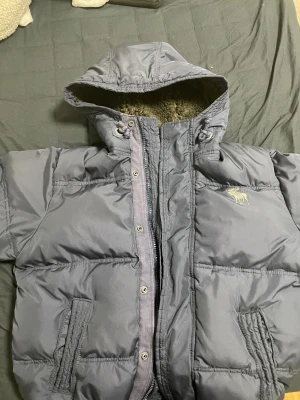 Mörkblå pufferjacka Abercrombie S - Säljer en mörkblå pufferjacka från Abercrombie & Fitch i storlek S. Jackan har huva med mysigt teddyfoder, dragkedja och knappar framtill samt två fickor. Klassisk logga på bröstet och elastisk nederkant. Perfekt för kalla dagar.