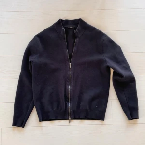 Svart stickad kofta med dragkedja från Massimo Dutti - Svart stickad kofta från Massimo Dutti med hel dragkedja framtill och ribbad krage. Tröjan har långärmade smala ärmar och är tillverkad i mjuk ull som känns skön mot huden. Perfekt för dig som gillar stilrena och enkla plagg med snygg passform. Storleken är L men passar lite mer som M