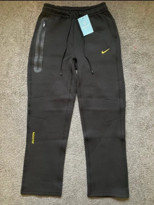 Svarta NOCTA mjukisbyxor från Nike - Svarta mjukisbyxor från Nike NOCTA med gul logga och detaljer. Byxorna har snörning i midjan, dragkedja på fickan och tryckta bokstäver på benen. Materialet är mjukt och bekvämt, perfekt för chill eller träning. Snygg och modern design med raka ben. Använda en gång 
