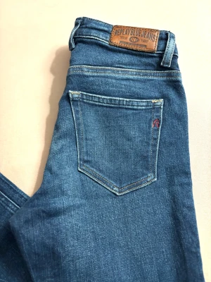 Replay Katewin blå jeans, storlek 25 - Snygga blå jeans från Replay, modell Katewin, med klassisk femficksdesign och brunt läderpatch bak i midjan. Jeansen har raka ben och normal passform, tillverkade i mjukt denim-material med subtila slitningar. Perfekt för dig som gillar stilrena och tidlösa jeans.