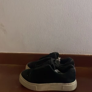 Svarta Eytys skor - Svarta slip-on sneakers med tjock vit platt sula och elastisk ovandel. Skorna har en enkel design utan snörning och en mörklila detalj på hälen. Materialet ser ut att vara syntet eller textil, perfekt för dig som gillar stilrena och bekväma skor.