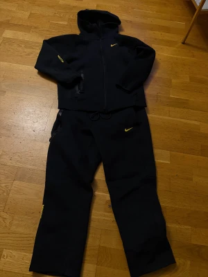 Svarta NOCTA x Nike dress - Svarta dress från Nike NOCTA med gul logga på benet och dragsko i midjan. Byxorna har raka ben, sidofickor med dragkedja och är gjorda i mjukt bomullsmaterial. Perfekt för chill eller träning, riktigt clean design med coola detaljer.