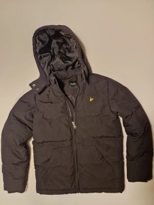 Svart vinter pufferjacka från Lyle & Scott Nypris 1400kr - Svart pufferjacka från Lyle & Scott med huva och dragkedja framtill. Jackan har det klassiska gula logobroderiet på bröstet och två fickor framtill. Perfekt för kalla dagar och har en stilren, enkel design. Nypris: 1400kr Pris kan diskuteras.
