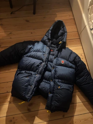 Blå dunjacka från Fjällräven med huva - Säljer en blå dunjacka från Fjällräven med huva, gul snörning och dragkedja framtill. Jackan har två stora fickor med knappar och Fjällräven-logga på bröstet. Perfekt för kalla dagar och har en riktigt schysst oversized look. Den är storlek S och tror att det antingen är man eller unisex. Säljer då jag själv köpte denna second hand och trodde det var dam. Den är i väldigt bra skick utan fläckar eller missfärgning! Pris kan diskuteras men går gärna inte mycket lägre 