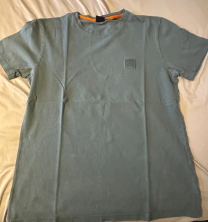 Grön kortärmad t-shirt från BOSS - Snygg grön t-shirt från BOSS med korta ärmar och rund hals. T-shirten har en diskret BOSS-logga broderad på bröstet och en orange detalj på insidan av kragen. Tillverkad i mjuk bomull för skön känsla.