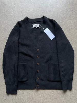 Svart kofta från Maison Margiela - Svart ribbstickad kofta från Maison Margiela med v-ringning, bruna knappar och två framfickor. Ikoniska vita stygn i nacken. Långärmad och tillverkad i mjukt ullmaterial. Perfekt för lager-på-lager och stilrena outfits.