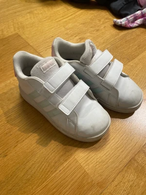 Vita adidas sneakers med kardborre - Säljer ett par vita adidas sneakers med klassiska tre ränder i ljusblått på sidan. Skorna har två breda kardborreband för enkel på- och avtagning, rund tå och platt sula med hexagonmönster undertill. Ovandelen är i syntetmaterial och mesh för skön komfort.