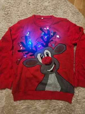 Röd jultröja med renmotiv och lampor - Rolig röd stickad jultröja i storlek M med ett stort renmotiv på framsidan, där nosen är en fluffig röd boll och hornen är dekorerade med blinkande lampor. Perfekt för att sticka ut på julfirandet! Rund hals och långa ärmar.