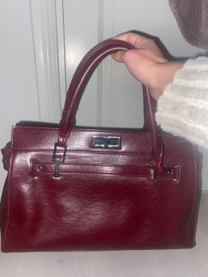 Vinröd handväska från Bershka - Snygg vinröd handväska från Bershka i skinnimitation med stilrena silverdetaljer och dubbla handtag. Väskan har en klassisk, boxig form och ett rymligt innerfack med dragkedja. Perfekt för dig som gillar att ha med dig allt du behöver och vill ha en chic look.