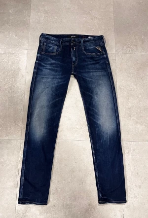 Replay Anbass jeans mörkblå 32 - Säljer ett par Replay Anbass jeans i mörkblå tvätt med snygga slitningar och klassiska fem fickor. Jeansen har gul kontrastsöm, rak passform och är tillverkade i slitstarkt bomullsmaterial. Perfekt för dig som gillar en stilren och modern look.