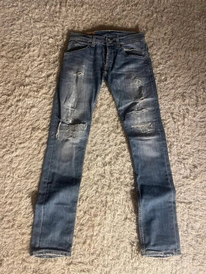Dondup jeans - Säljer ett par blå jeans från Dondup med slitna detaljer och trasiga partier på benen. Modellen har klassisk femficksdesign, dragkedja vid bakfickan och snygga slitningar för en cool look. Perfekt för dig som gillar streetstyle och vill sticka ut.
