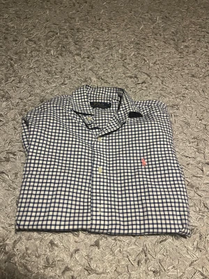 Rutig skjorta från Polo Ralph Lauren - Säljer en vit och blå rutig skjorta från Polo Ralph Lauren i slim fit. Skjortan har klassisk krage, långa ärmar och knappar framtill. Materialet är mjuk bomull och den har den ikoniska rosa Polo-loggan broderad på bröstet.
