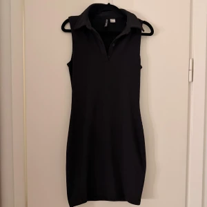 Svart ribbad kortklänning  - Svart ärmlös kortklänning från H&M Divided med ribbad struktur och krage. Klänningen har v-ringning med knappar framtill och en slim passform som framhäver siluetten. Perfekt för dig som gillar stilrena och enkla plagg med en sportig touch.