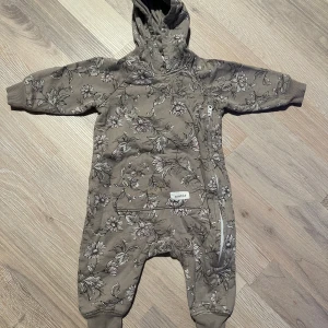 Blommig beige babyoverall från Newbie - Säljer en beige babyoverall från Newbie med blommigt mönster i ljusa toner. Overallen har huva, ribbade muddar och en praktisk dragkedja på sidan. Perfekt för mysiga dagar och enkel att ta på och av.