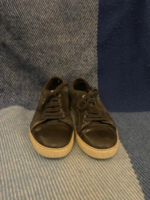 Svarta sneakers från Lanvin - Säljer ett par svarta sneakers från Lanvin med snygg mocka på sidorna och blank svart tå i skinn. Skorna har snörning och vit platt sula med Lanvin-logga undertill. Perfekt för dig som gillar stilrena och klassiska sneakers med exklusiv känsla.tyvär var inne sulan för förstörd så att behövde slänga kom med frågor 