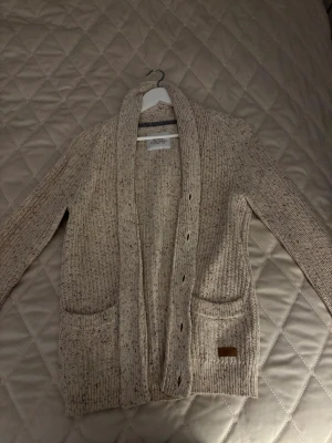 Beige stickad kofta - Säljer en beige stickad kofta med grov ribbstickning och mörka knappar framtill. Koftan har två praktiska fickor och en liten Lexington-logga på ena fickan. Perfekt för lager-på-lager och chill dagar. Aldrig använd