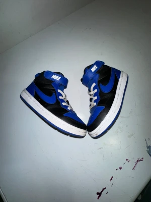 Nike Jordan 1 Mid Blå/Svart Barn - Säljer ett par Nike Jordan 1 Mid i blått och svart med vita detaljer. Skorna har klassisk snörning, kardborreband upptill och vit sula med blå undersida. Ikonisk Nike Swoosh på sidorna och hög modell som ger en sportig vibe. Perfekta för dig som gillar streetstyle.