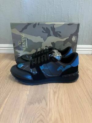 Valentino Rockrunner sneakers blå/svart - Snygga Valentino Rockrunner sneakers i svart och blått med coolt camouflage-mönster. Skorna har platt sula med nitar, rund tå och snörning. Materialmix av skinn och mocka ger en lyxig känsla. Perfekta för dig som vill sticka ut med stil. Strlk 45 men passar egentligen 44