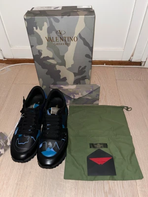 Valentino Garavani Rockrunner sneakers - Säljer ett par Valentino Garavani Rockrunner sneakers i svart och blått med coolt kamouflagemönster. Skorna har snörning, platt sula med grovt mönster och detaljer i skinn och textil. Kommer med originalkartong, dustbag och taggar.