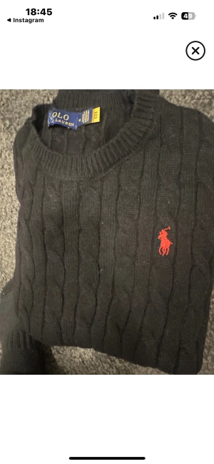 Svart kabelstickad tröja Polo Ralph Lauren - Svart kabelstickad tröja från Polo Ralph Lauren med klassisk röd logobrodering på bröstet. Rund halsringning och ribbade muddar. Perfekt för dig som gillar stilrena och tidlösa plagg med snygga detaljer.