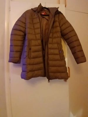 Brun dunjacka från Luhta med huva - Säljer en brun dunjacka från Luhta i storlek 36. Jackan har huva med snörning, quiltad design och dragkedja framtill. Perfekt för kalla dagar och har en snygg, lite längre passform. Fickor med knappstängning på sidorna. Riktigt chill och varm jacka! Dragkedjan är ganska jobbig att fp igen men är rätt så.säker på att man bara kan olja den.