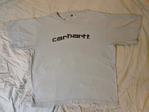 Ljusblå Carhartt t-shirt med logga - Snygg ljusblå t-shirt från Carhartt med svart logotyp tryckt på bröstet. Klassisk rund halsringning och korta ärmar. Tillverkad i mjuk bomull som är skön mot huden. Mindre missfärgning på framsidan ( se sista bilden ) men det synns knappt. 