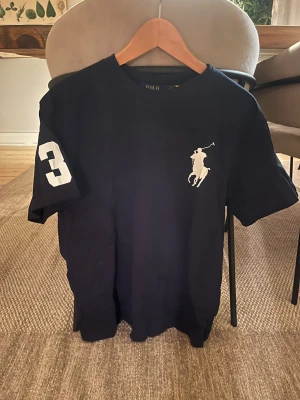 Navy t-shirt från Polo Ralph Lauren - Navy t-shirt från Polo Ralph Lauren med stort vitt polospelar-tryck på bröstet och siffran 3 på ärmen. Klassisk rund hals och normal passform. Perfekt för dig som gillar sportig och stilren look.