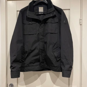 Svart fieldjacket från J.Lindeberg - Svart fieldjacket från J.Lindeberg med cool patch på ärmen och stora fickor med knappar framtill. Jackan har klassisk krage, knappstängning och är långärmad. Perfekt för dig som gillar stilren och funktionell design.