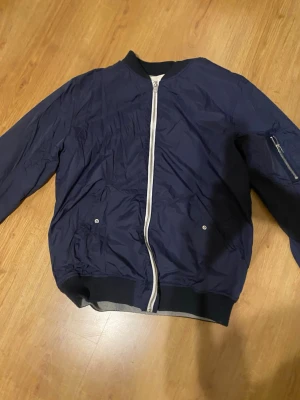 Mörkblå bomberjacka från L.O.G.G. - Snygg mörkblå bomberjacka från L.O.G.G. med vit dragkedja framtill och ribbade muddar vid ärmslut och nederkant. Jackan har en ficka med dragkedja på ärmen och två sidofickor med tryckknappar. Perfekt för dig som gillar stilren och enkel design.