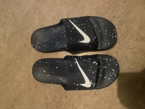 Svarta Nike sandaler med vit splatter - Säljer ett par svarta Nike sandaler med vit splattermönster och stor vit Swoosh-logga på ovansidan. Sandalerna har en unik, upphöjd sula med mönstrad undersida för extra komfort. Perfekta för chill dagar och enkel att dra på.