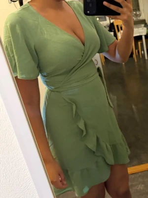 Grön omlottklänning med volang - Snygg ljusgrön omlottklänning med korta ärmar och volangdetaljer längs nederkanten. Klänningen har v-ringad hals och knyt i midjan för en smickrande passform. Perfekt för dig som gillar en feminin och trendig stil.
