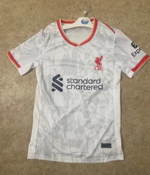 Vit Liverpool FC matchtröja - Säljer en vit Liverpool FC matchtröja med subtilt rutigt mönster, röda och gula detaljer vid krage och ärmslut. Tröjan har klubbens logga i rött på bröstet och sponsortryck i mörkblått. Perfekt för dig som älskar fotboll och vill visa ditt stöd.
