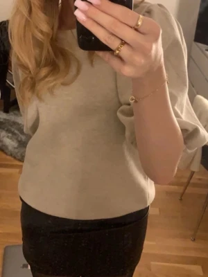Beige blus med puffärmar - Säljer en stilren beige blus med puffiga trekvartsärmar och rund halsringning. Blusen har en enkel och elegant design med lite små glitter i tyget, som passar perfekt till både jeans och kjol. Materialet känns mjukt och bekvämt mot huden.