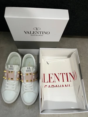Valentino Rockstud Untitled sneakers - Säljer ett par vita Valentino Rockstud Untitled sneakers med ljusrosa band och guldiga nitar över ovansidan. Skorna har snörning, rund tå och platt sula. Materialet är skinn och detaljerna ger en lyxig vibe. Kommer med originalkartong och dustbag. Passar dig som vill ha en lyxig stil.