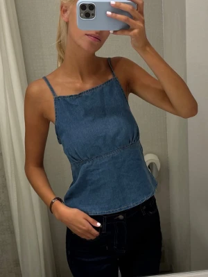 Blå denim linnetopp med smala axelband - Säljer en blå linnetopp i denim med smala justerbara axelband och rak halsringning. Toppen har en lätt utsvängd passform och synliga sömmar som ger en cool look. Perfekt för varma dagar och enkel att matcha med jeans eller shorts.
