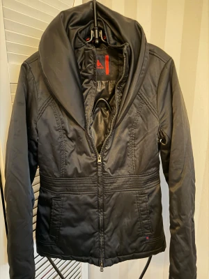 Svart pufferjacka från Robe di Kappa - Svart pufferjacka från Robe di Kappa med bred krage och snygga sömdetaljer runt midjan. Jackan har dragkedja framtill, två fickor och långärmade ärmar. Materialet är glansigt och känns som polyester, perfekt för kyliga dagar. Stilren och enkel design med diskret logga.