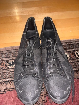 Svarta sneakers från Gucci med GG-mönster - Säljer ett par svarta Gucci sneakers med klassiskt GG-mönster över hela skon. Skorna har rund tå, platt sula och snörning. Materialet är en mix av skinn och textil, vilket ger en lyxig känsla. Perfekt för dig som gillar exklusiva märken och stilren design.