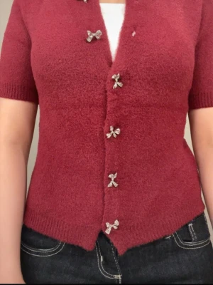 Vinröd kofta med rosettknappar - Superfin vinröd kofta med korta ärmar och fluffigt stickat material. Snygga silverfärgade rosettknappar framtill ger en gullig touch. Perfekt att styla med jeans eller kjol för en mysig look. Rund halsringning och ribbade muddar vid ärmslut och nederkant.