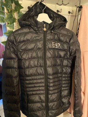 Svart pufferjacka från Emporio Armani - Notera att detta är AI text: Svart pufferjacka från Emporio Armani EA7 med huva och dragkedja framtill. Jackan har ett quiltat mönster och logotypen i vitt på bröstet. Materialet är glansigt och känns lätt, perfekt för kyliga dagar. EA7 jacka, 2 år sedan köp. Användt få gånger då jag inte gillar halskragar. Original och äkta är den finns kod du kan scanna på bilderna köpt för ca 1600