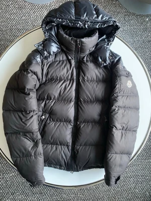 Moncler Maya  - En mycket fin moncler Maya. Den är matsvart men glansig på axlarna och huvan. Mycket fint skick några defekter vilket syns på bilderna. Hör gärna av dig kring funderingar. Den är stolen L men passar M. 