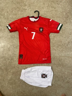 Portugal fotbollströja Ronaldo #7 - Säljer en röd Portugal fotbollströja med nummer 7 och Ronaldo på ryggen, samt vita shorts med Portugal-emblem. Tröjan har små vita mönster, gröna och vita detaljer på ärmarna och är gjord i polyester med DryCell-teknologi. Perfekt för dig som älskar fotboll och vill ha en snygg matchoutfit.