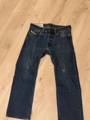 Blå jeans från Diesel, storlek 31 - Säljer ett par klassiska blå jeans från Diesel i storlek 31. Jeansen har rak passform, fem fickor och snygga detaljer med Diesel-logga på bakfickan. Tillverkade i 100% denim med italiensk design. Perfekt för dig som gillar tidlös stil.