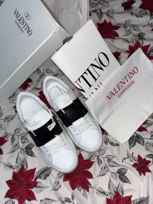 Valentino Garavani Open sneakers vit/svart - Säljer ett par Valentino Garavani Open sneakers i vit och svart. Skorna har en bred svart läderdetalj över mitten, rund tå och vita snören. Märkeslogga i guld på plösen. Kommer med originalkartong och dustbag. Riktigt snygga och stilrena sneakers i skinn.