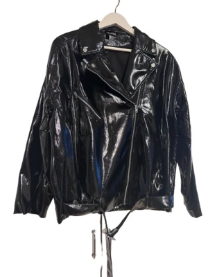 Svart glansig skinnjacka med bälte - Säljer en svart glansig skinnjacka med klassisk bikerstil, sned dragkedja och coolt bälte i midjan. Jackan har långa ärmar, bred krage med tryckknappar och en riktigt edgy look. Perfekt för dig som vill sticka ut med en statement piece.
