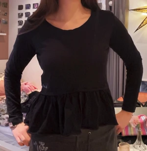 Svart volangblus från Bondelid - Svart långärmad blus från Bondelid med rund halsringning och volang nedtill. Tillverkad i mjuk bomull och har en enkel, stilren design med liten broderad logga framtill. Perfekt att styla med jeans eller kjol.