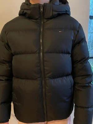 Svart vinterjacka från Tommy Hilfiger - Fin svart vinterjacka från Tommy Hilfiger strl 164 (passar en xs/s). Den är köpt från Kids Brand Store. Endast använd fåtal gånger.                            Köpte den för 2300kr. 